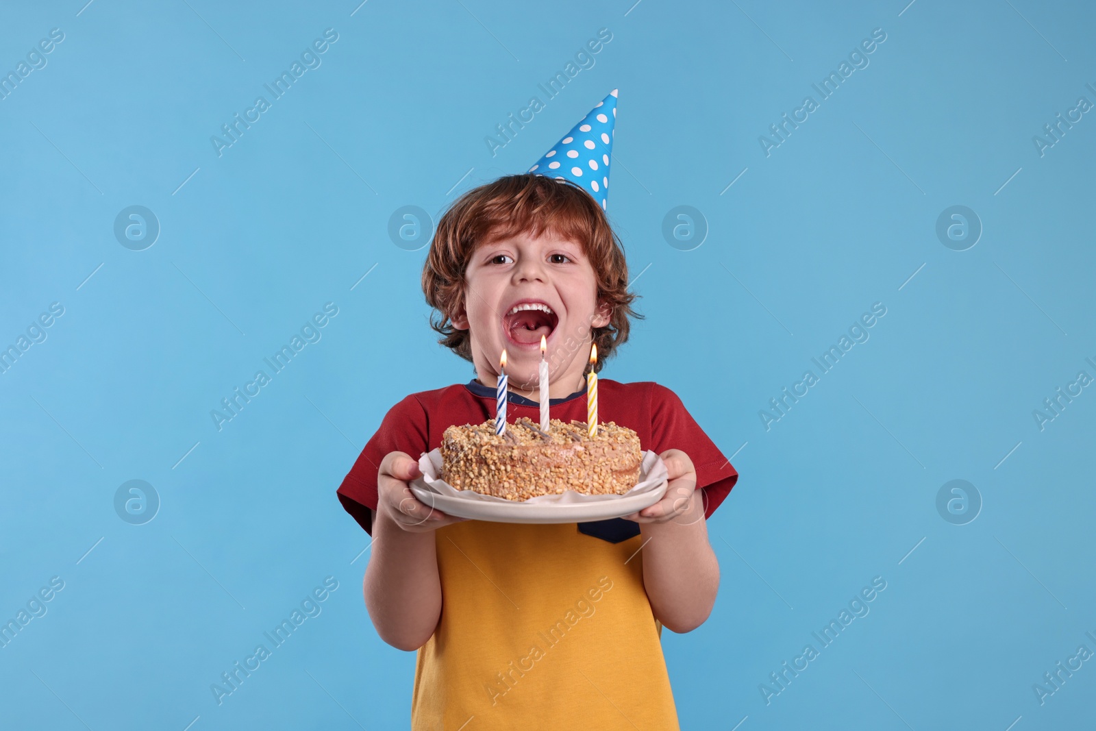 Photo of Birthday celebration. Cute little boy in party hat holding tasty cake with burning candles on light blue background