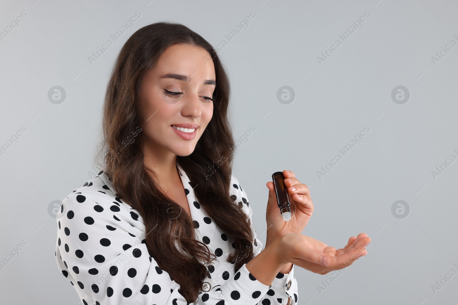 Photo of Beautiful happy woman with roller bottle applying essential oil onto wrist on light grey background