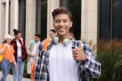 Photo of Students spending time together outdoors. Happy young man with notebooks showing thumbs up, selective focus