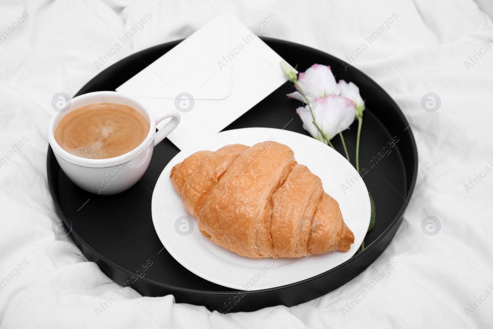 Photo of Tray with tasty croissant, cup of coffee and flowers on white bed