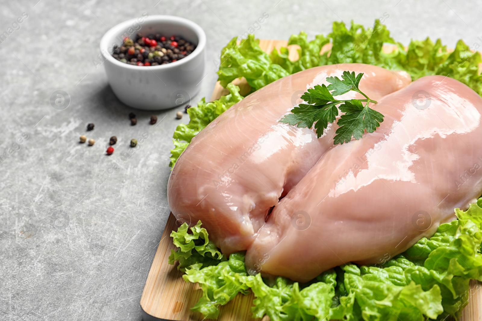 Photo of Wooden board with raw chicken breasts and lettuce near peppers mix in bowl on grey background