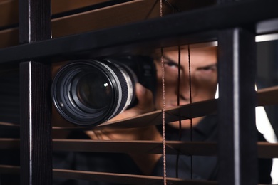 Photo of Private detective with camera spying near window indoors, focus on lens