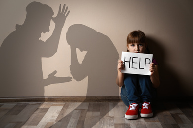 Image of Sad little girl with sign HELP sitting on floor and silhouettes of arguing parents 