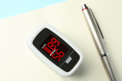 Photo of Modern fingertip pulse oximeter, pen and open notebook on light blue background, flat lay