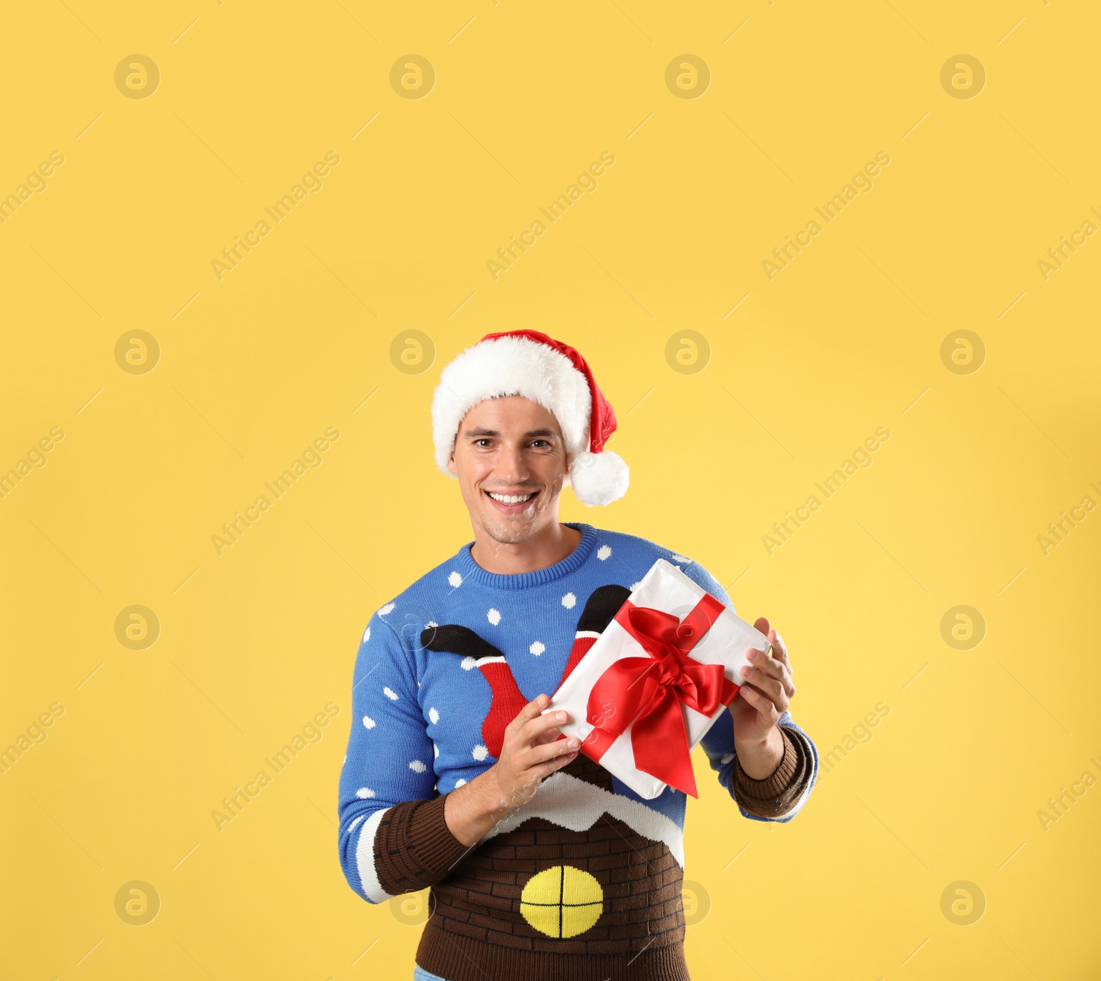 Photo of Happy man in Christmas sweater and Santa hat holding gift box on yellow background