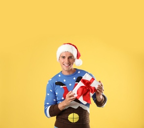 Happy man in Christmas sweater and Santa hat holding gift box on yellow background