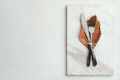 Cutlery, dry leaf and marble board on white wooden background, top view with space for text. Table setting elements