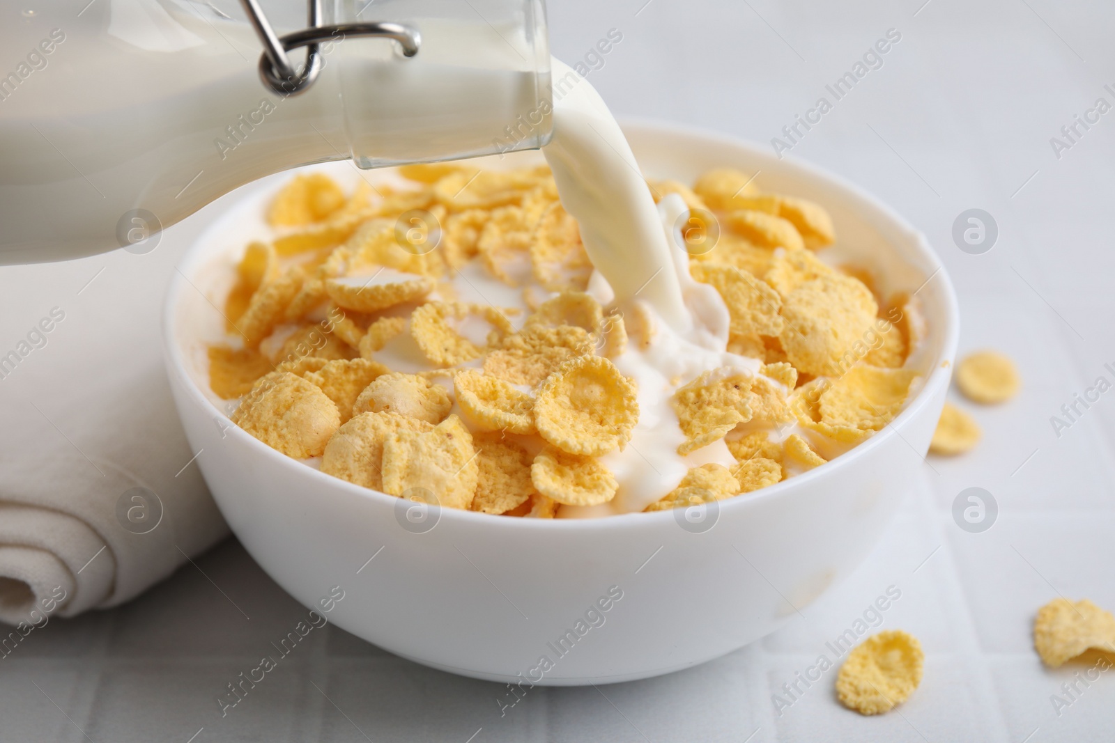 Photo of Breakfast cereal. Pouring milk into bowl with tasty corn flakes at white table, closeup