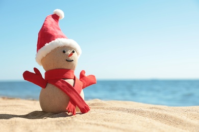 Photo of Snowman made of sand with Santa hat and scarf on beach near sea, space for text. Christmas vacation