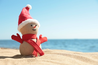 Snowman made of sand with Santa hat and scarf on beach near sea, space for text. Christmas vacation