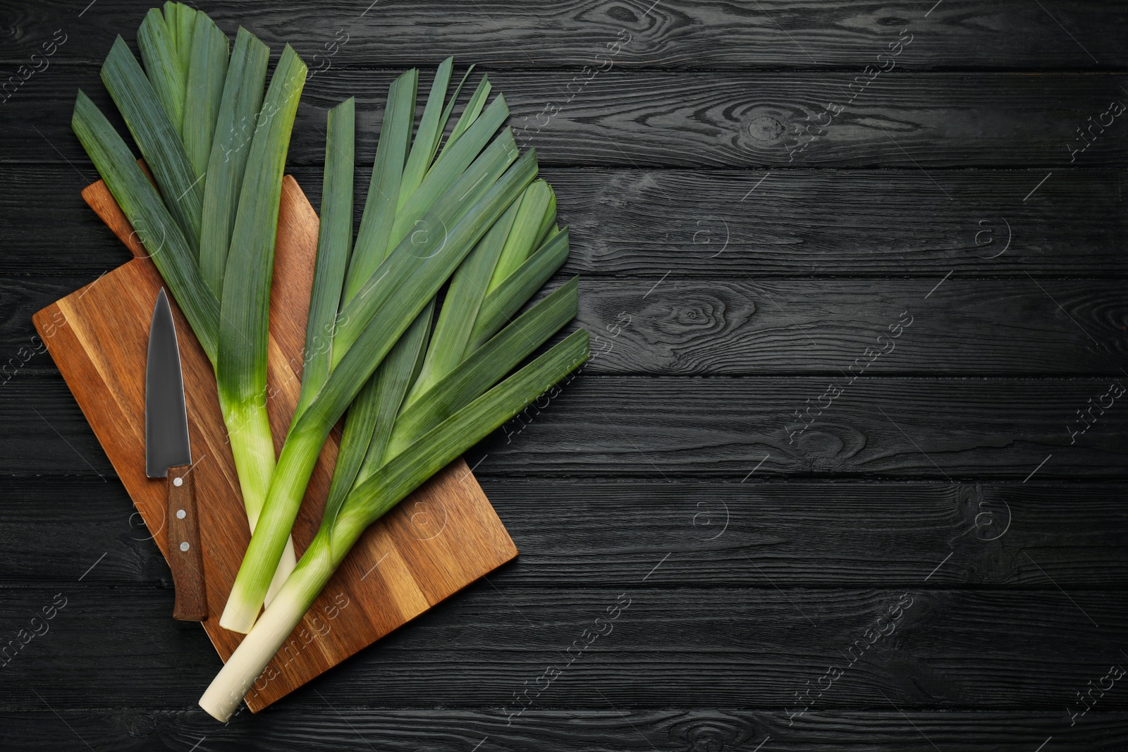 Photo of Fresh raw leeks and knife on black wooden table, top view. Space for text