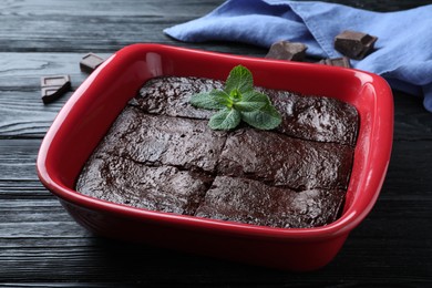 Photo of Delicious chocolate brownie with mint in baking dish on black wooden table