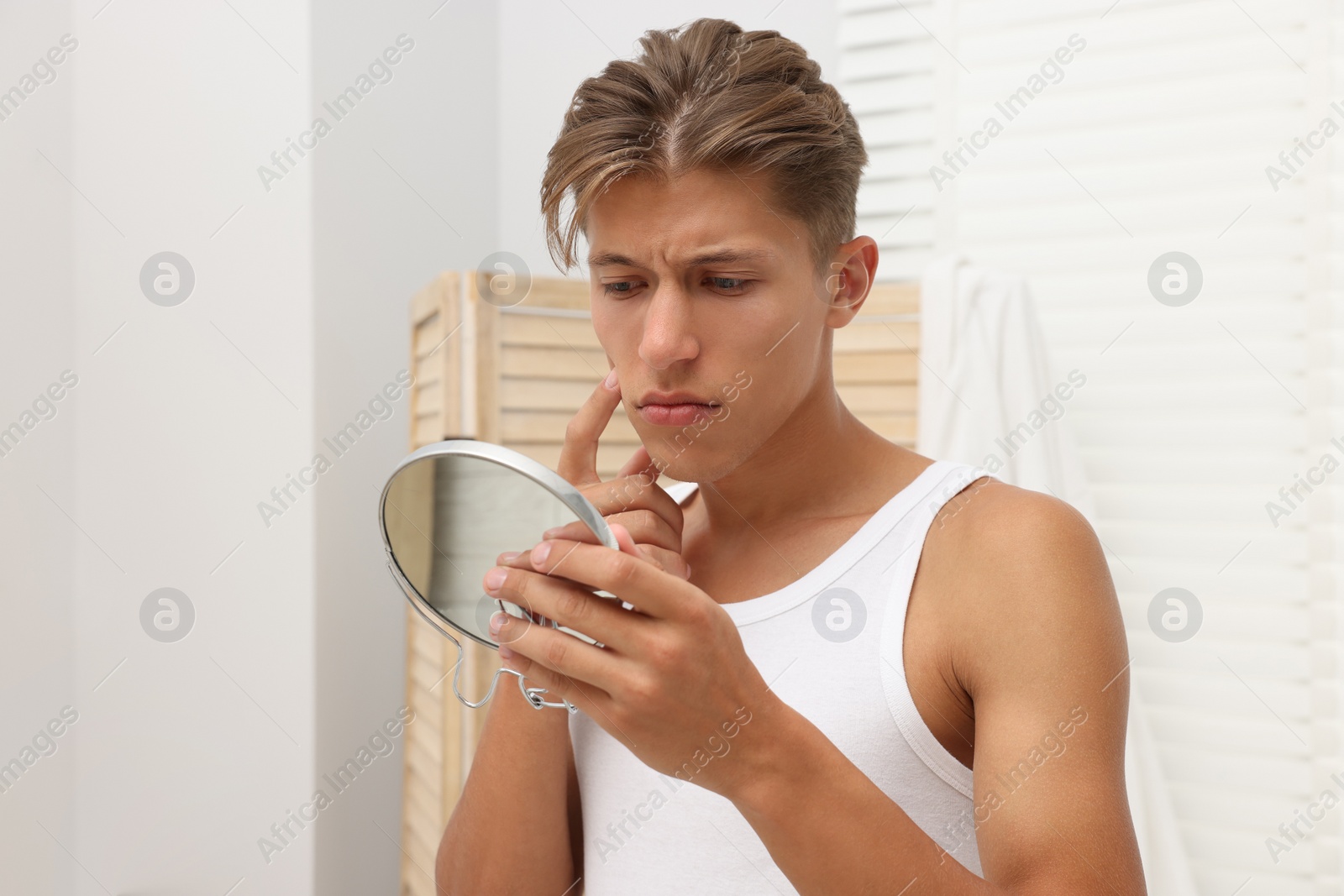 Photo of Upset young man looking at mirror and touching pimple on his face indoors. Acne problem