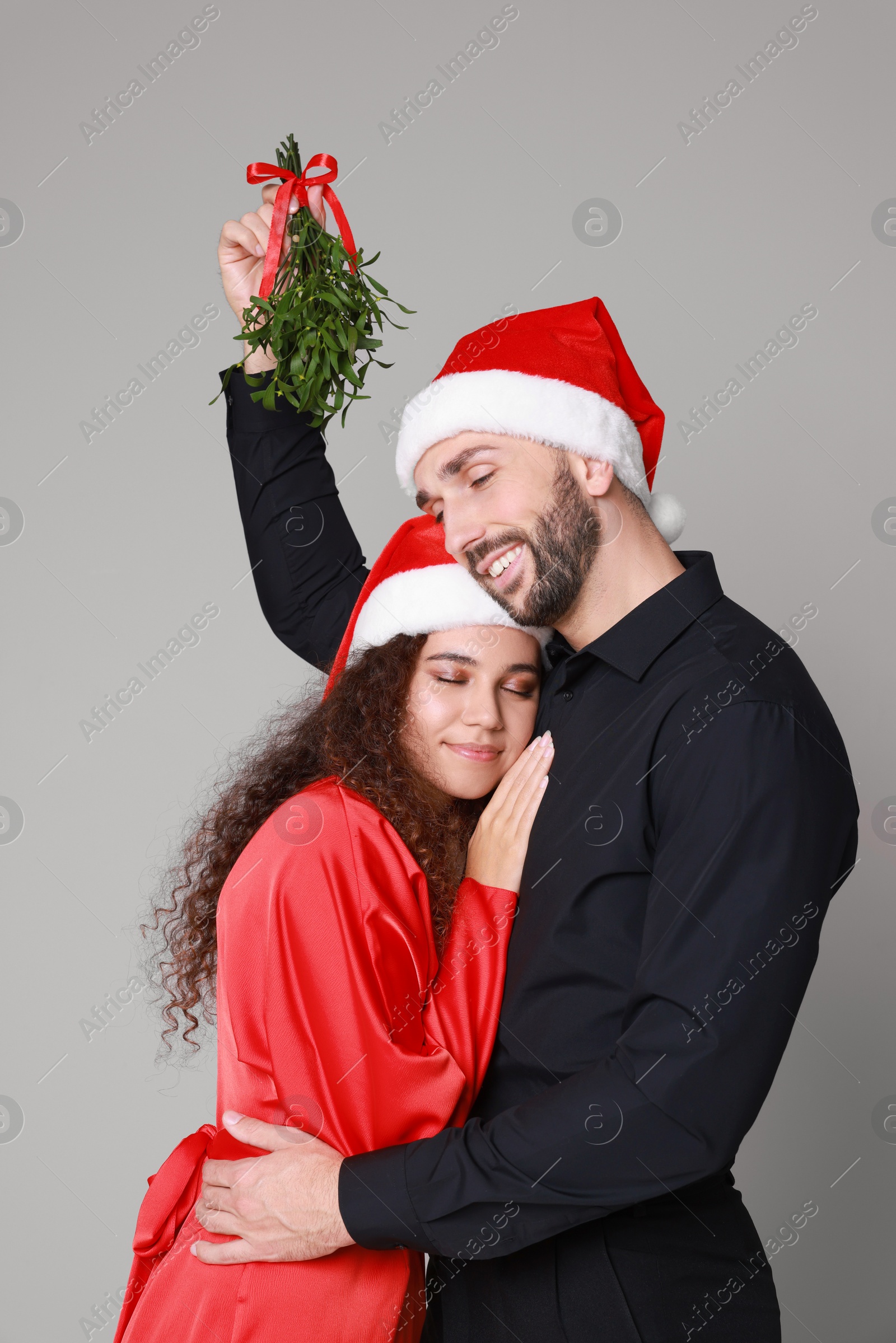 Photo of Lovely couple in Santa hats under mistletoe bunch on grey background