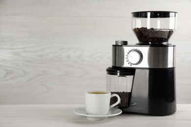 Photo of Modern electric coffee grinder and cup of fresh drink on white wooden table. Space for text