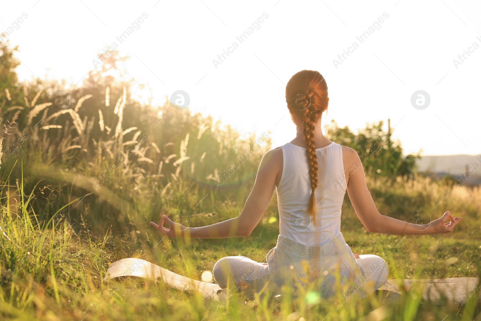 Photo of Woman practicing Padmasana on yoga mat outdoors on sunny day, back view. Lotus pose