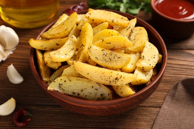 Photo of Bowl with tasty baked potato wedges, spices and garlic on wooden table, closeup