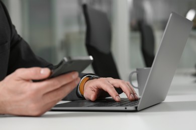 Photo of Man using phone while working on laptop at white desk in office, closeup