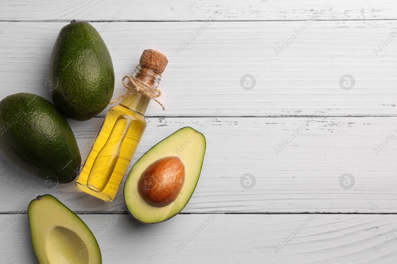 Photo of Glass bottle of cooking oil and fresh avocados on white wooden table, flat lay. Space for text