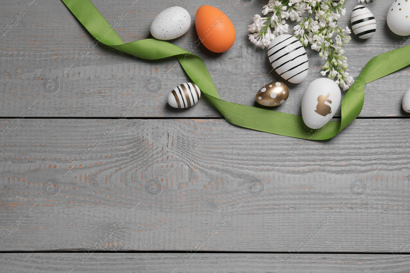 Photo of Many painted Easter eggs, branch with lilac flowers and ribbon on grey wooden table, flat lay. Space for text