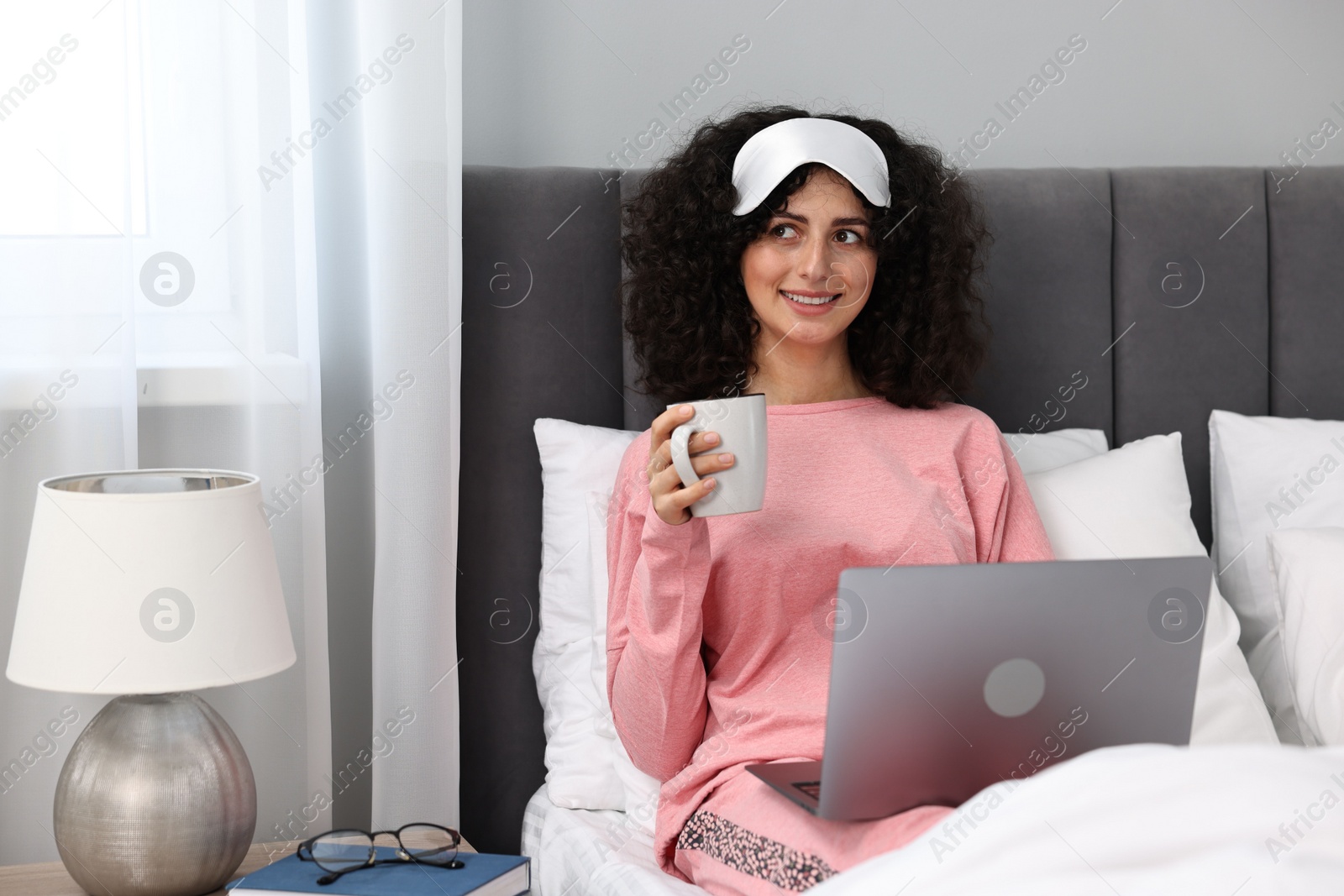 Photo of Beautiful young woman in stylish pyjama and sleep mask with cup of drink using laptop in bed at home