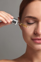 Beautiful young woman applying cosmetic serum onto her face on grey background, closeup