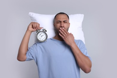Tired man with pillow and alarm clock yawning on light grey background. Insomnia problem