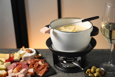 Photo of Fork with piece of ham, fondue pot with melted cheese, wine and snacks on grey table, closeup