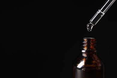 Dripping cosmetic serum from pipette into bottle on black background, closeup and space for text