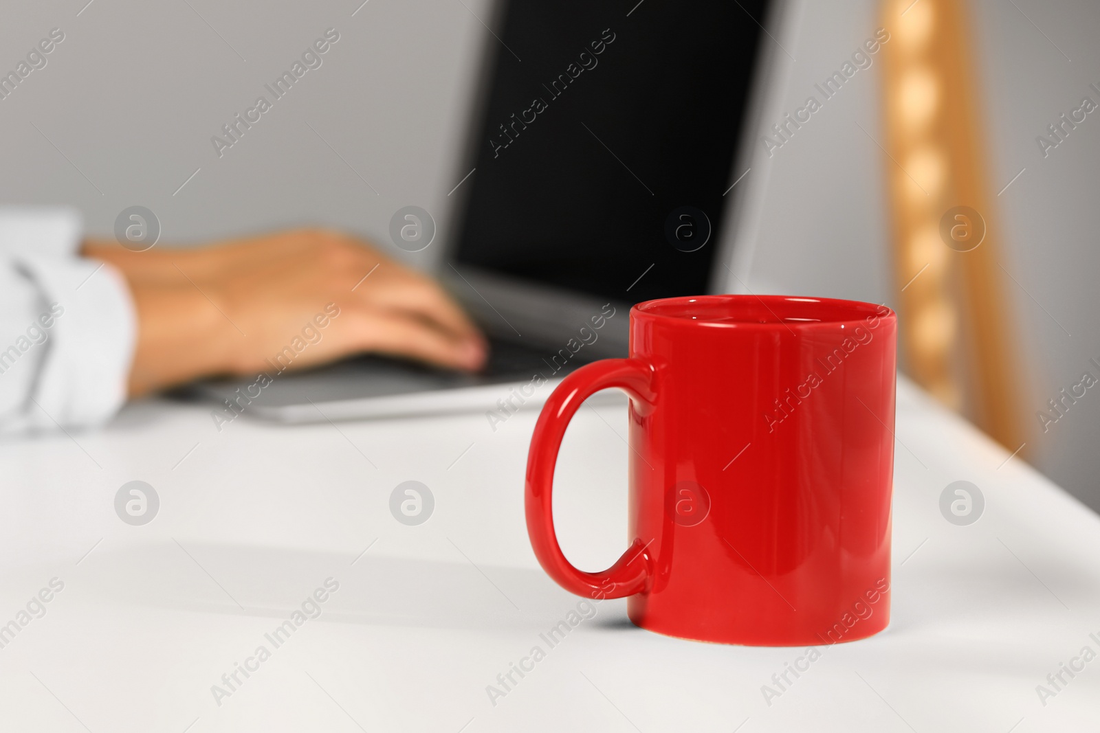 Photo of Red ceramic mug on white table, selective focus and space for text. Woman working with laptop at workplace, closeup