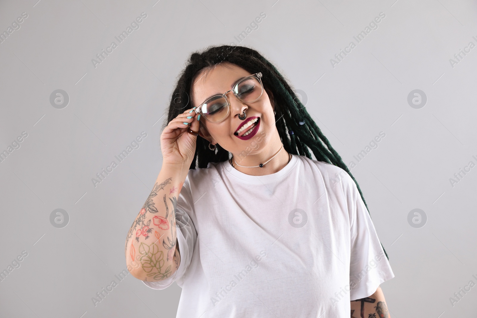 Photo of Beautiful young woman with tattoos on arms, nose piercing and dreadlocks against grey background