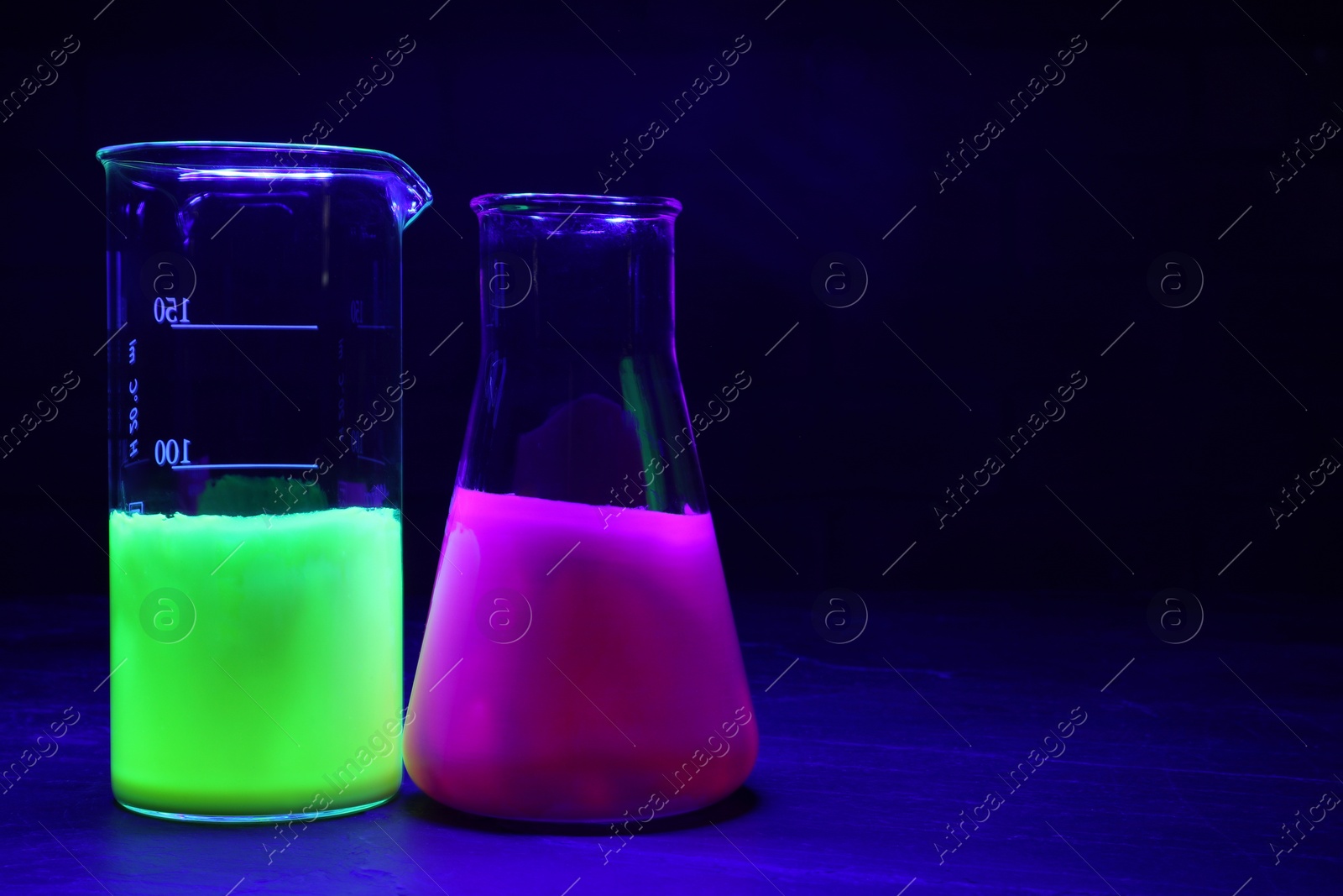 Photo of Laboratory glassware with luminous liquids on table against dark background, space for text