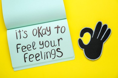 Photo of Notebook with phrase It`s Okay To Feel Your Feelings and paper cutout of OK hand gesture on yellow background, top view
