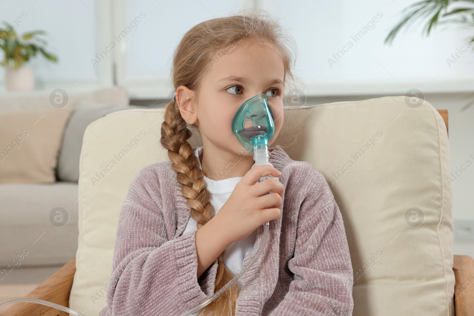 Photo of Little girl using nebulizer for inhalation in armchair at home