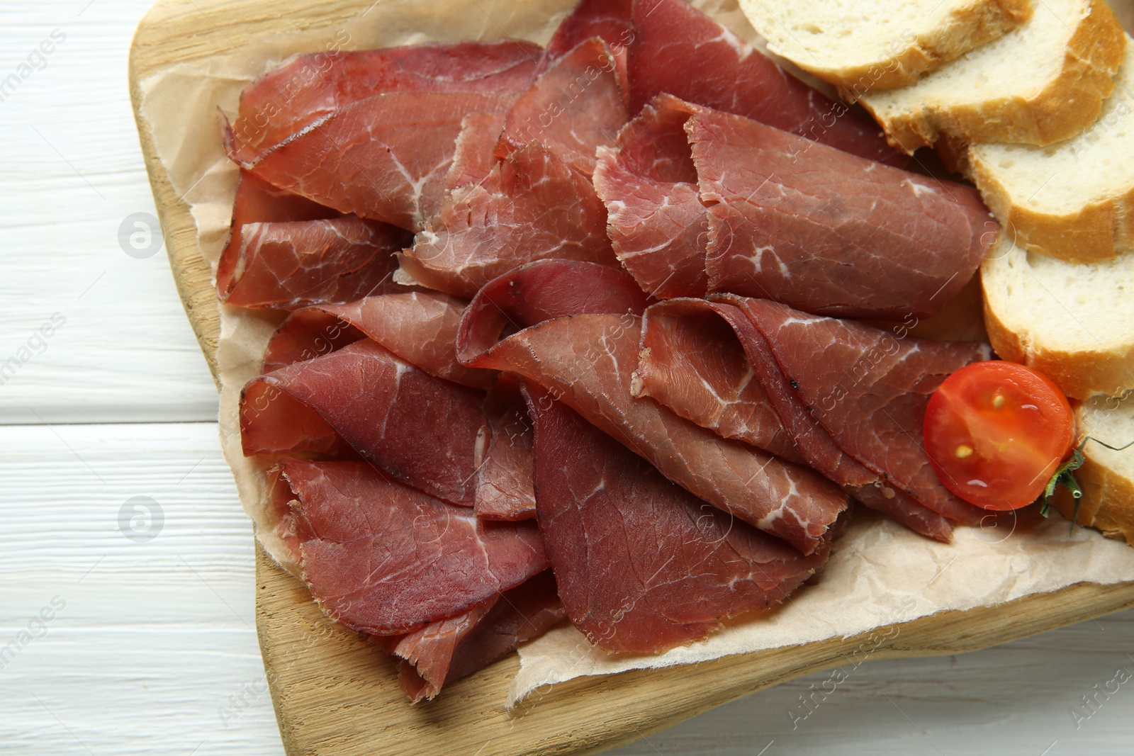 Photo of Board with delicious bresaola served with bread and tomato on white wooden table, top view