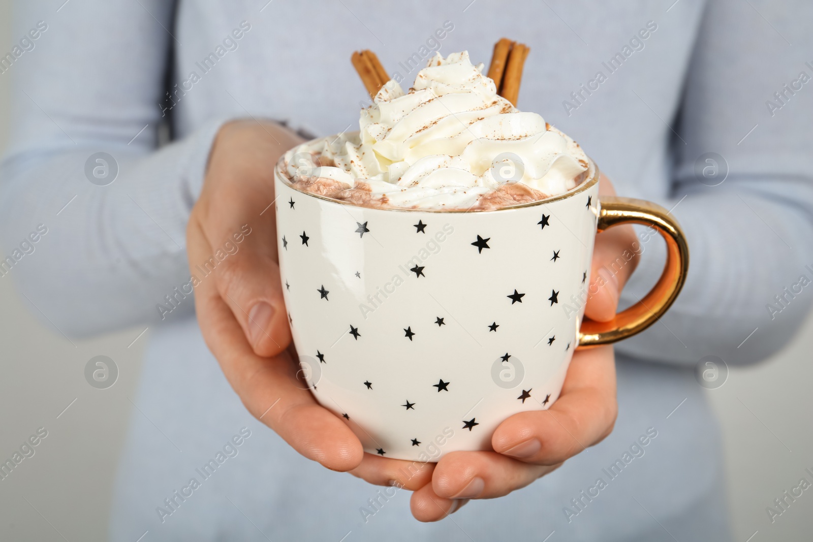 Photo of Woman holding cup of delicious hot chocolate with whipped cream and cinnamon sticks, closeup