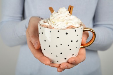 Photo of Woman holding cup of delicious hot chocolate with whipped cream and cinnamon sticks, closeup