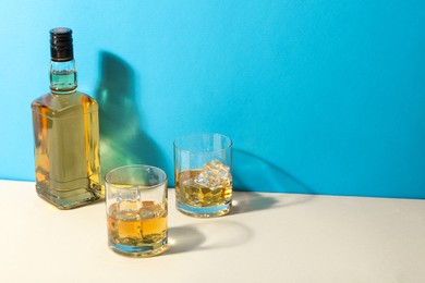 Whiskey with ice cubes in glasses and bottle on white table against light blue background, space for text