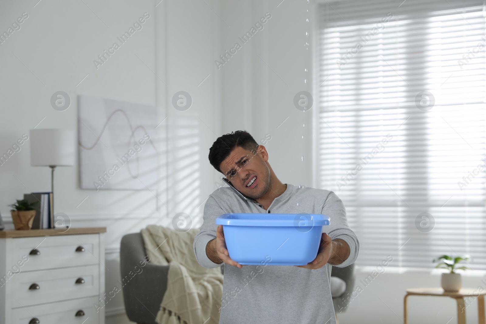 Photo of Emotional man calling roof repair service while collecting leaking water from ceiling in living room