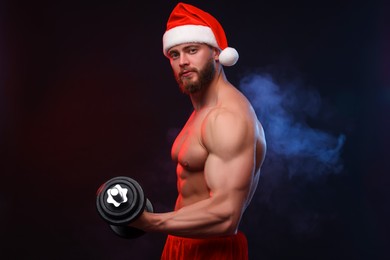 Photo of Muscular young man in Santa hat with dumbbell on black background