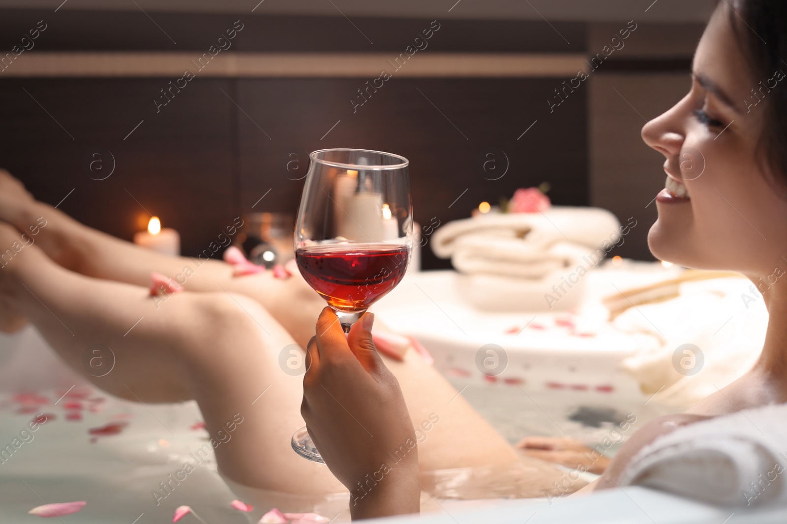 Photo of Woman holding glass of wine while taking bath with rose petals, closeup. Romantic atmosphere