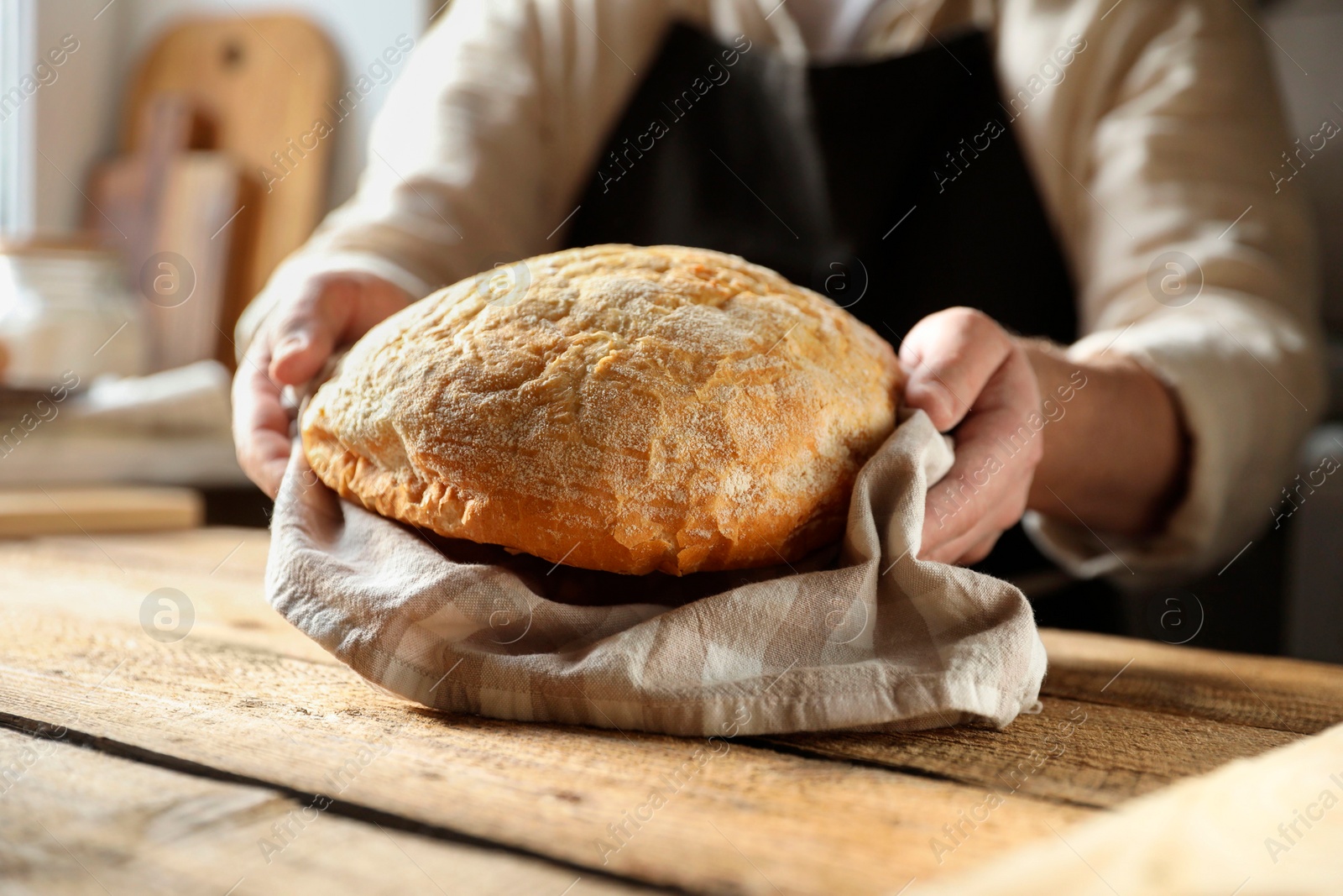 Photo of Man holding loaf of fresh bread at wooden table indoors, closeup