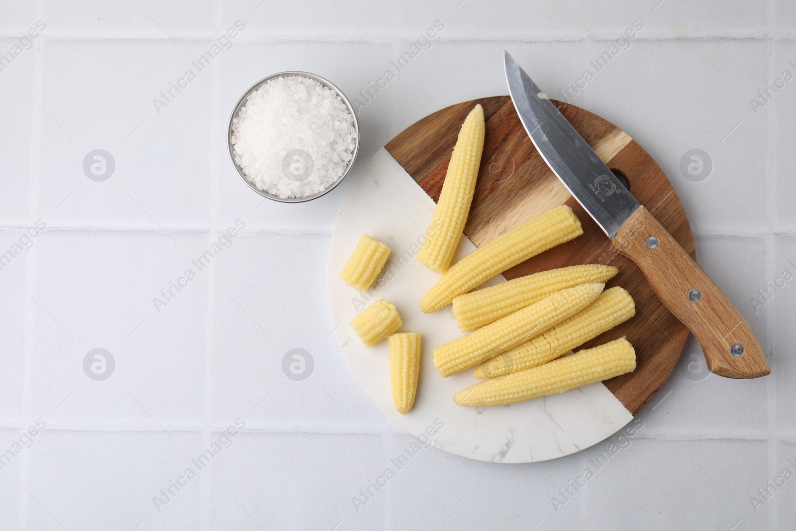 Photo of Tasty fresh yellow baby corns and knife on white tiled table, top view. Space for text