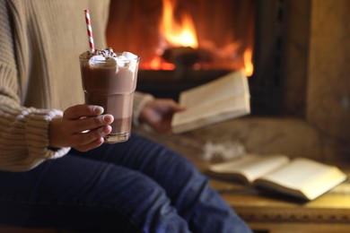 Woman with glass of hot cocoa and book near fireplace at home, closeup. Space for text