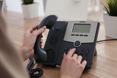 Photo of Woman using desktop telephone at wooden table in office, closeup. Hotline service