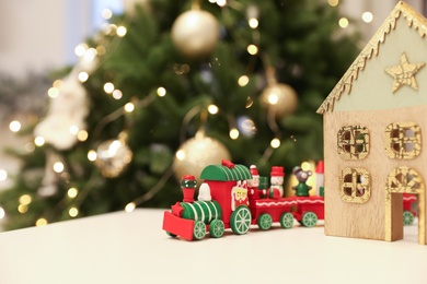 Photo of Bright toy train and decorative wooden house on white table near Christmas tree. Space for text