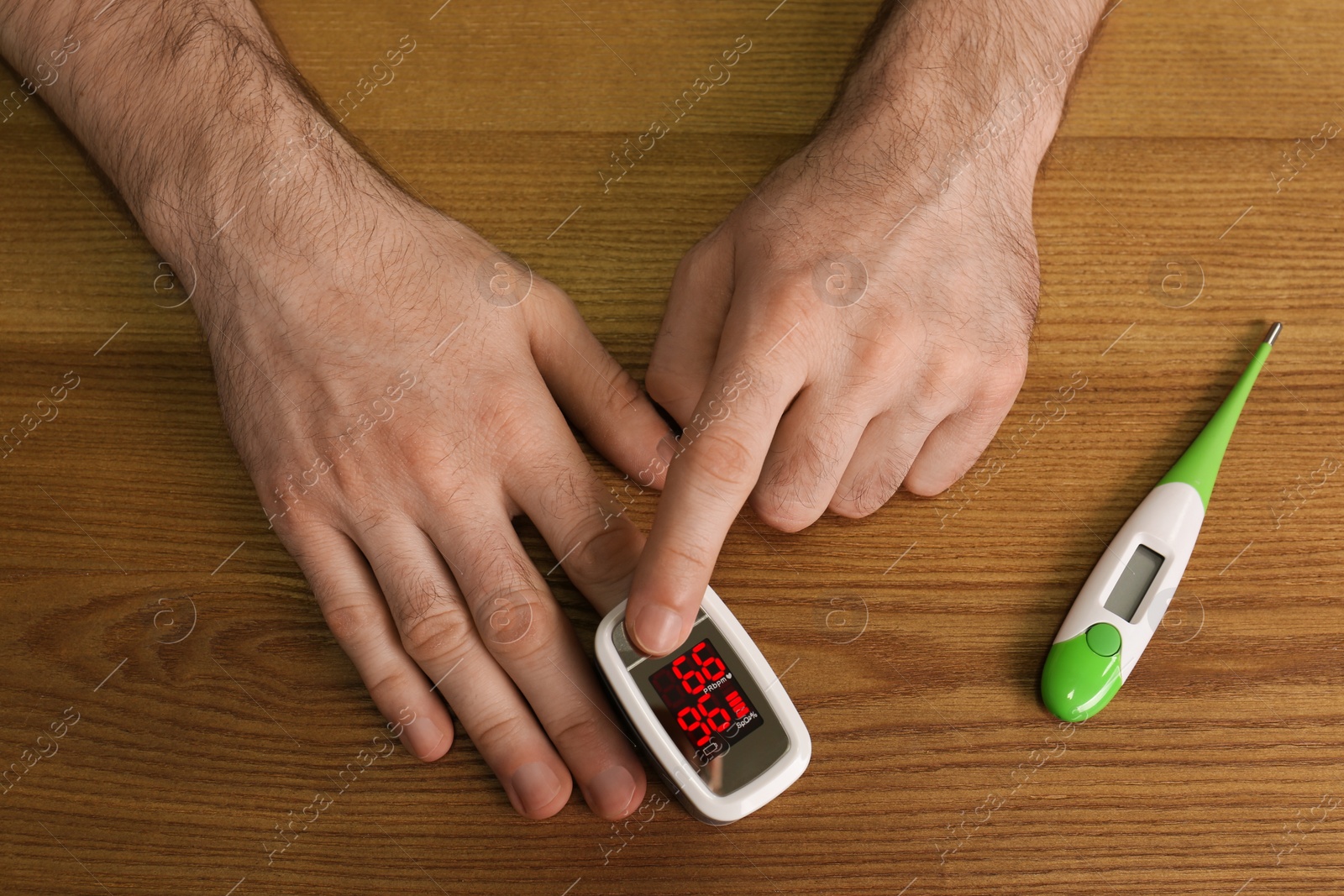 Photo of Man using pulse oximeter for oxygen level testing at wooden table with thermometer, top view