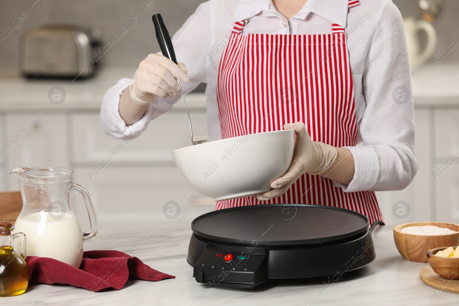 Photo of Woman cooking delicious crepe on electric pancake maker at white marble table in kitchen, closeup