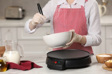 Woman cooking delicious crepe on electric pancake maker at white marble table in kitchen, closeup