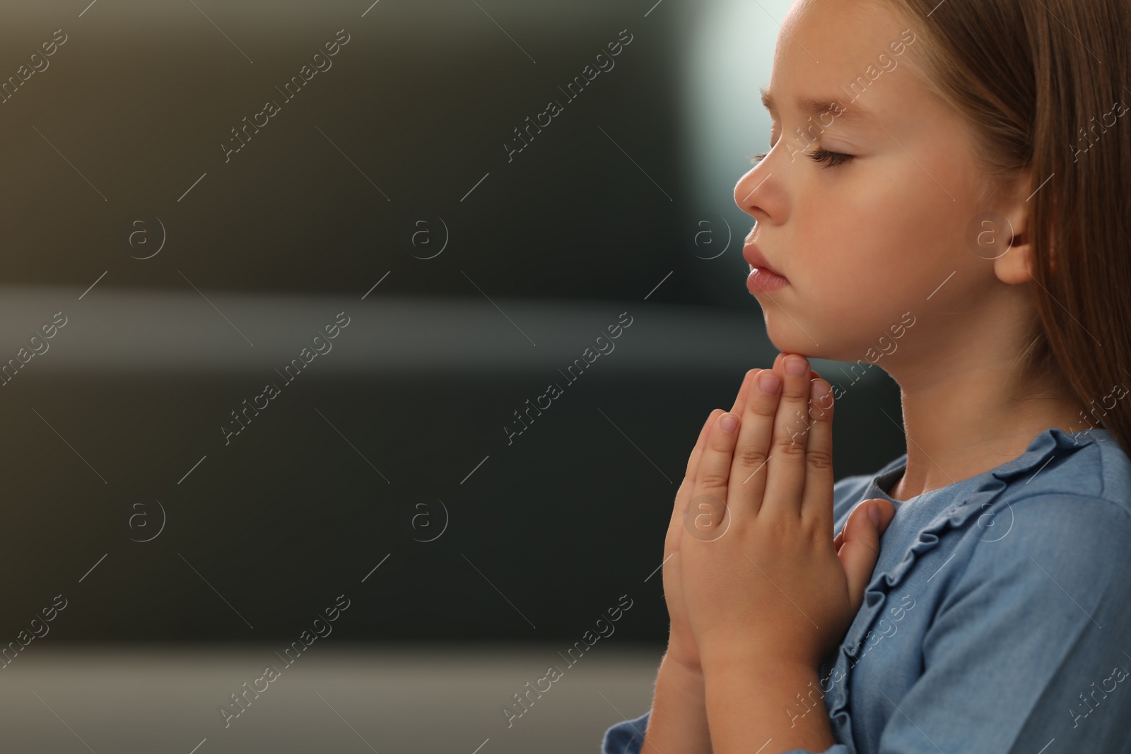 Photo of Cute little girl with hands clasped together praying on blurred background. Space for text